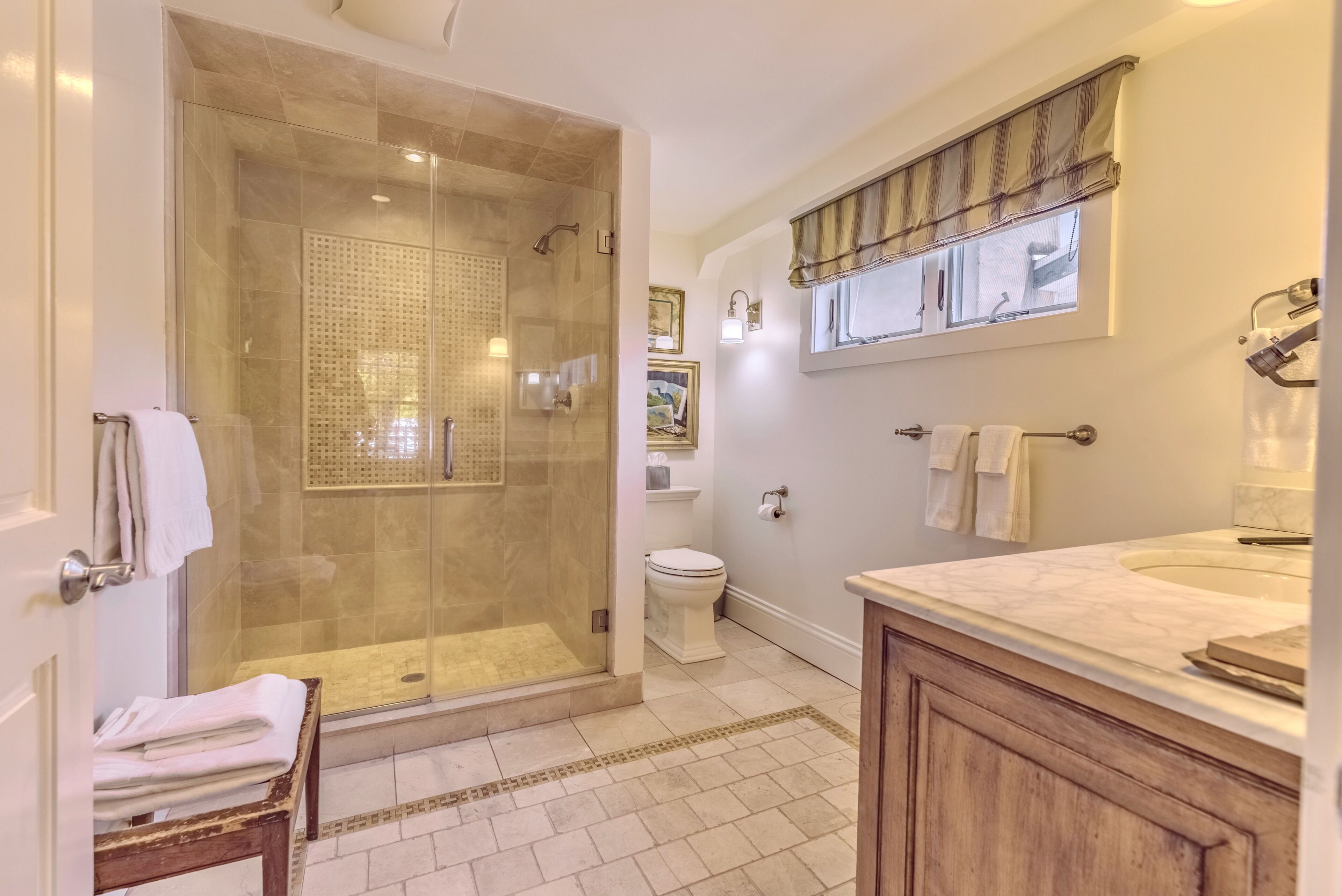 A bathroom with a glass-enclosed shower, a toilet, a sink with a marble countertop, and a window with curtains.