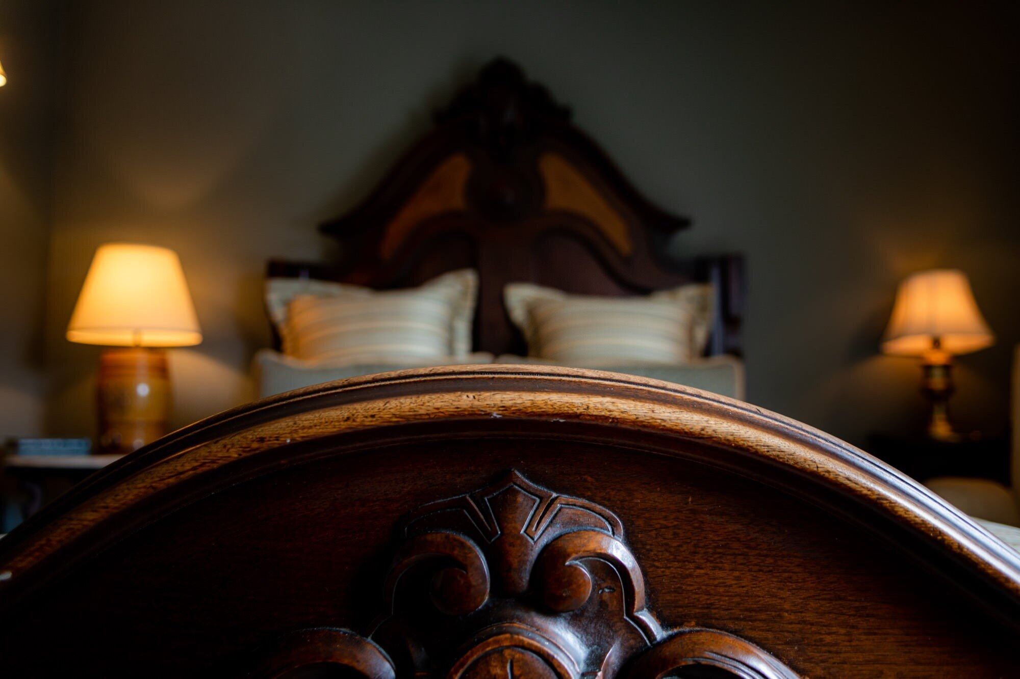 A close-up of a wooden headboard with intricate carvings, a bed with white linens, and two bedside lamps.