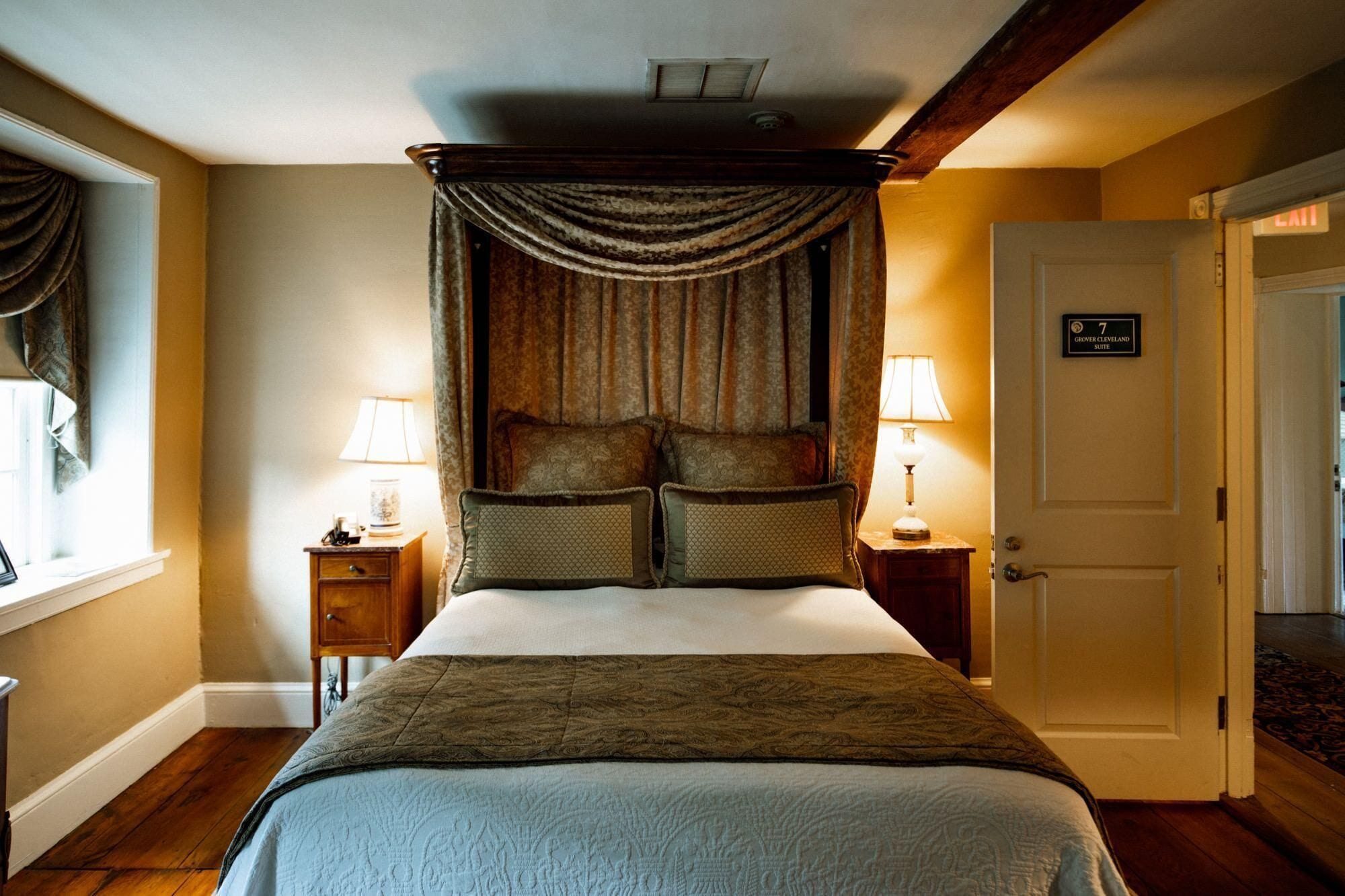 Quarto com cama de dossel, duas mesas de cabeceira com abajures, uma porta com número de suíte e uma janela com cortinas.