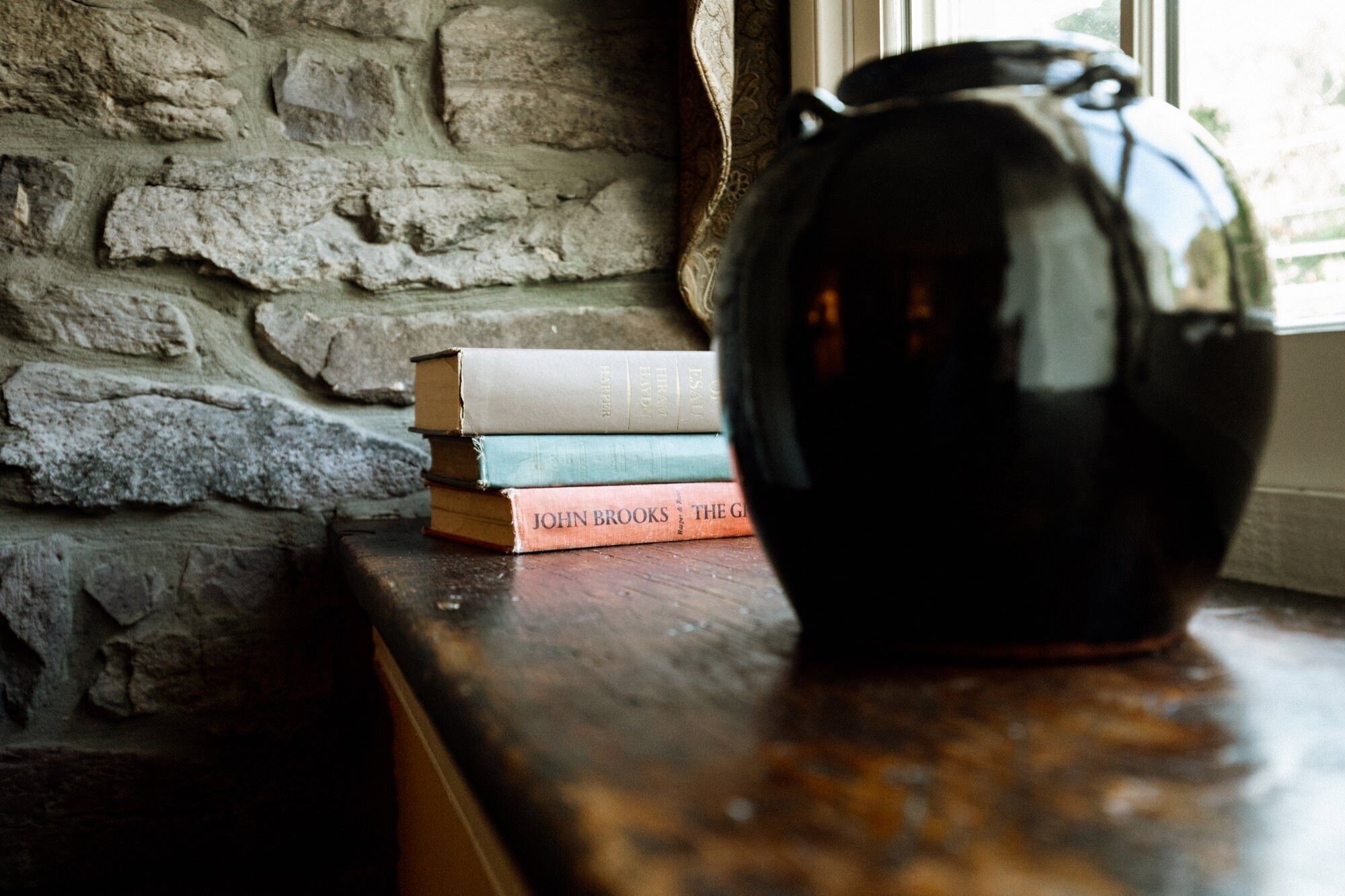 Un montón de libros sobre una superficie de madera, junto a un muro de piedra y un jarrón oscuro.