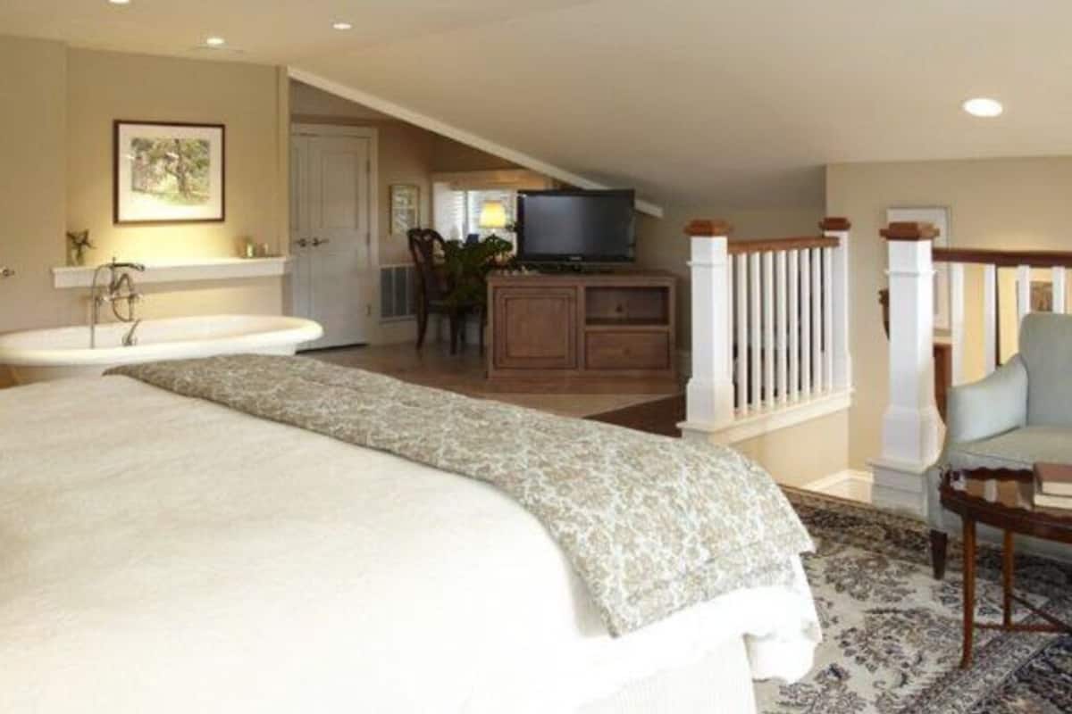 A bedroom with a large bed, a sitting area with a chair, a television, and a bathroom with a bathtub.