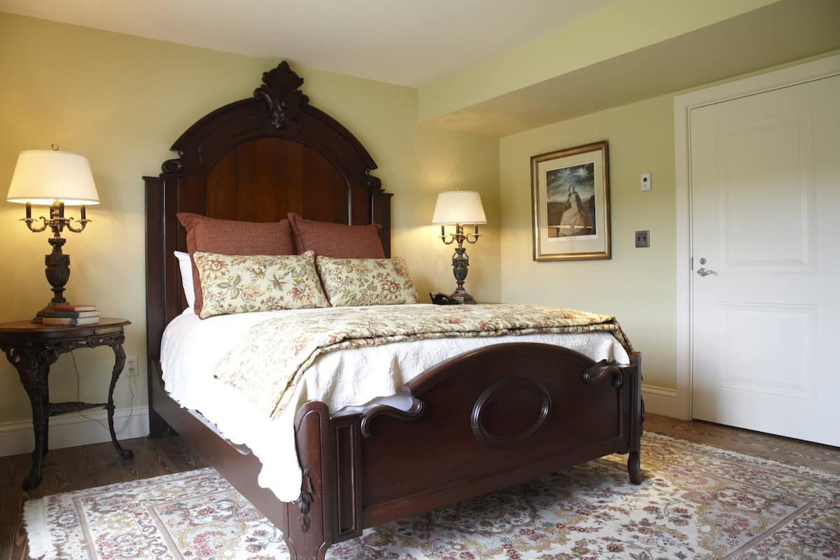 A bedroom with a wooden four-poster bed, two bedside tables with lamps, a floral-patterned rug, and a framed picture on the wall.