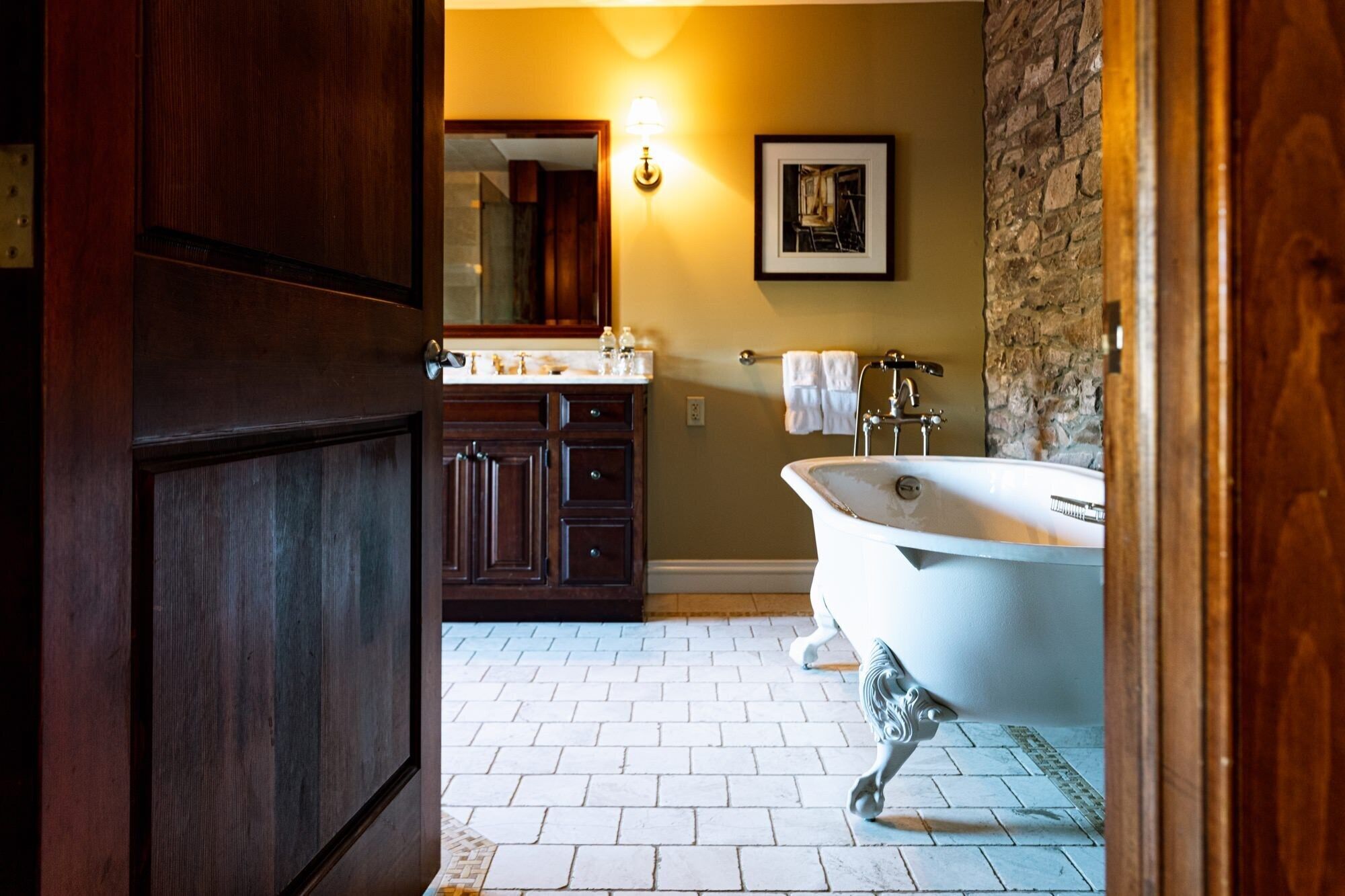 Une salle de bain avec une baignoire sur pieds, des meubles en bois, un mur d’accent en pierre et un sol carrelé.