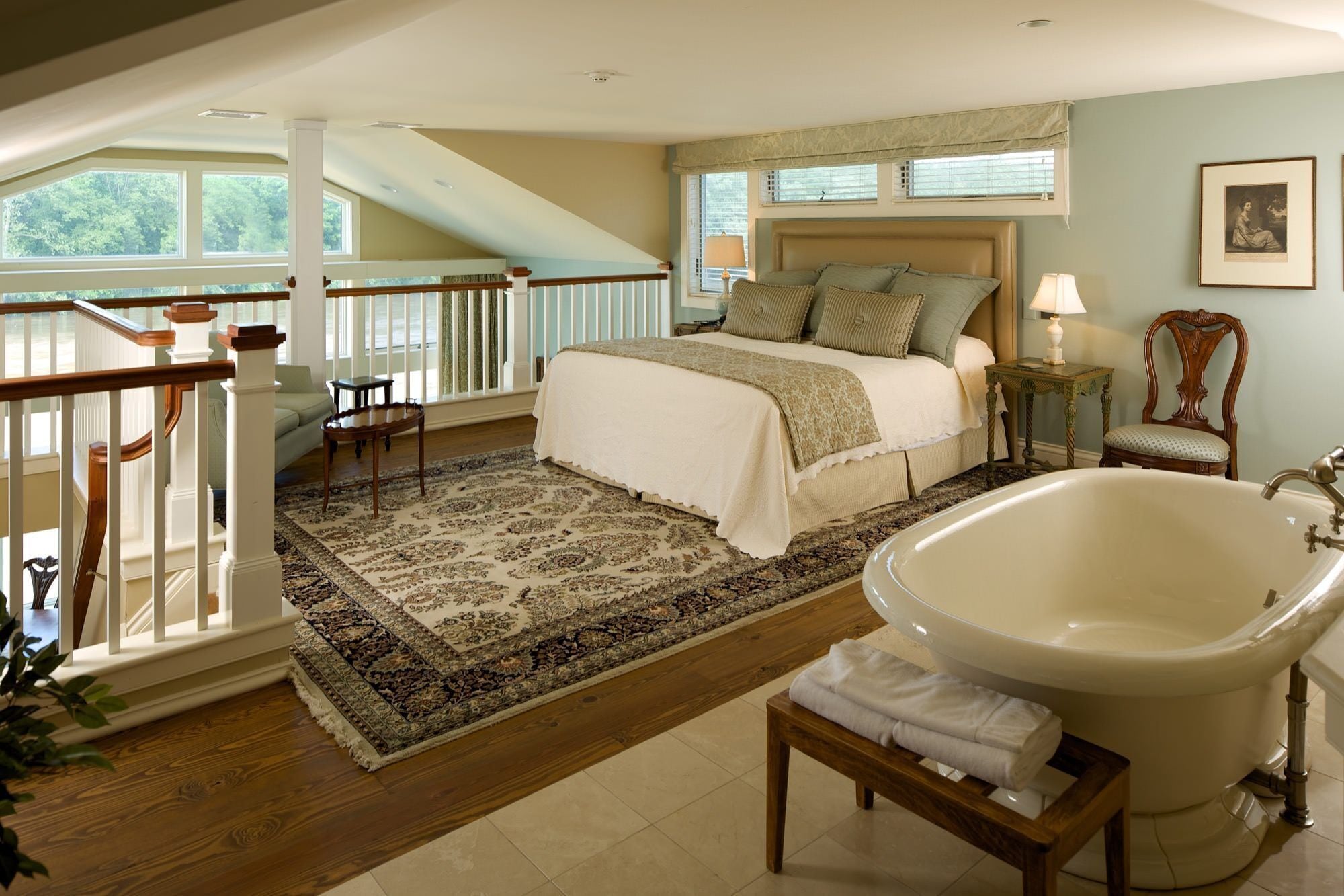 A spacious bedroom with a large bed, a sitting area with a chair, a wooden staircase, and a bathtub.