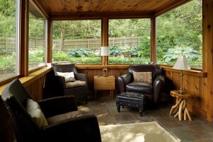A cozy room with wicker furniture, a leather armchair, and a view of a lush garden through large windows.