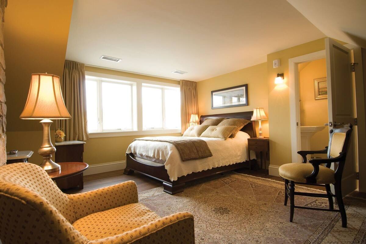 A spacious bedroom with a large bed, two bedside lamps, a chair, a small table, and a window with curtains.