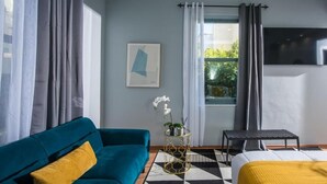Una habitación de hotel moderna con un sofá color turquesa, una cama con una manta amarilla, una mesita de noche con una planta, una silla negra y un ventanal con cortinas.