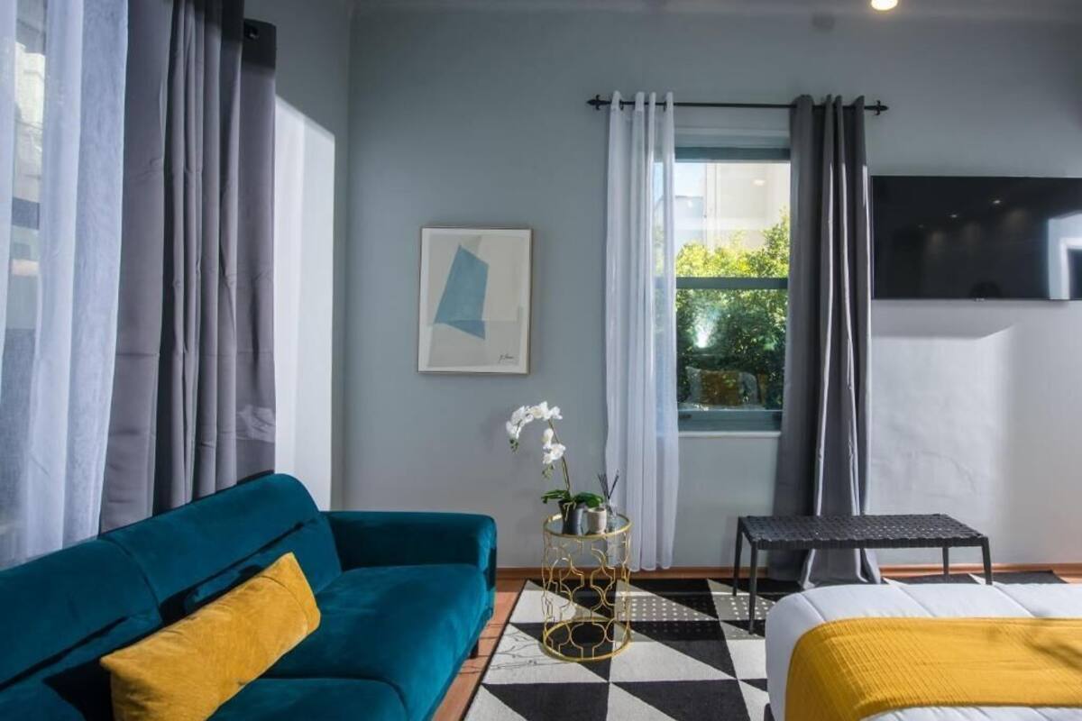 Ein modernes Hotelzimmer mit einem türkisfarbenen Sofa, einem Bett mit gelber Tagesdecke, einem kleinen Beistelltisch mit Pflanze, einem schwarzen Stuhl und einem großen Fenster mit Vorhängen.