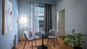 Una habitación con una mesa redonda, dos sillas, una lámpara de pie, una planta en maceta y un ventanal con cortinas.