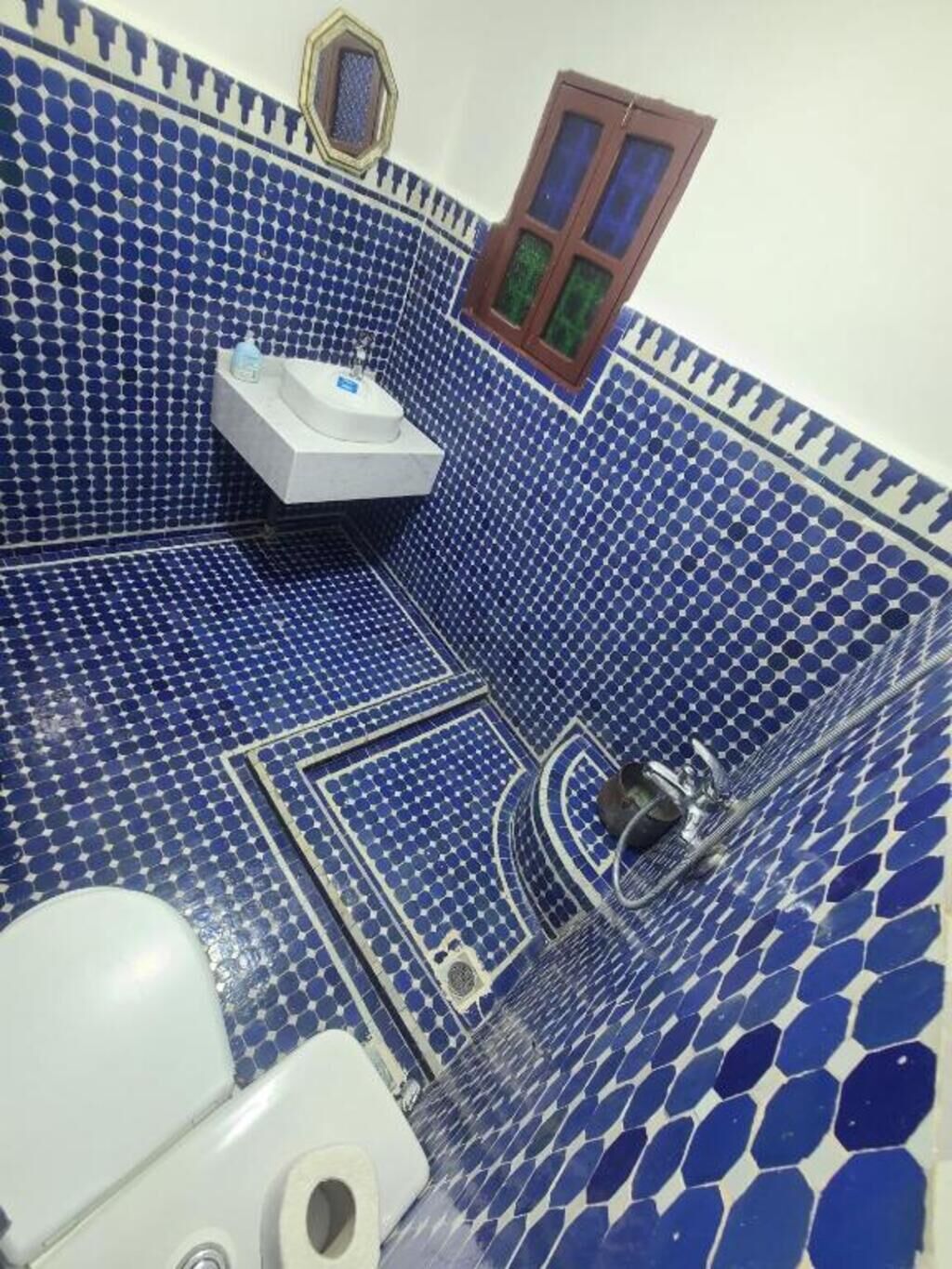 A bathroom with blue mosaic tiles, a white sink, a mirror, and a window with green curtains.