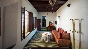 A room with a checkered floor, a sofa with red cushions, a hanging lamp, and a small table with a tablecloth. - Riad Lala Kenza (Fes)