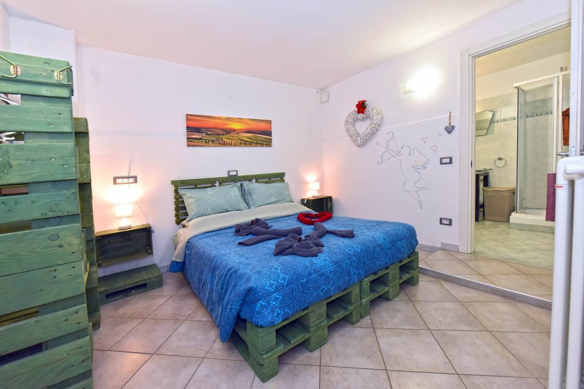 A bedroom with a bed, wooden nightstands, a painting, a heart-shaped decoration, and a view of the bathroom.