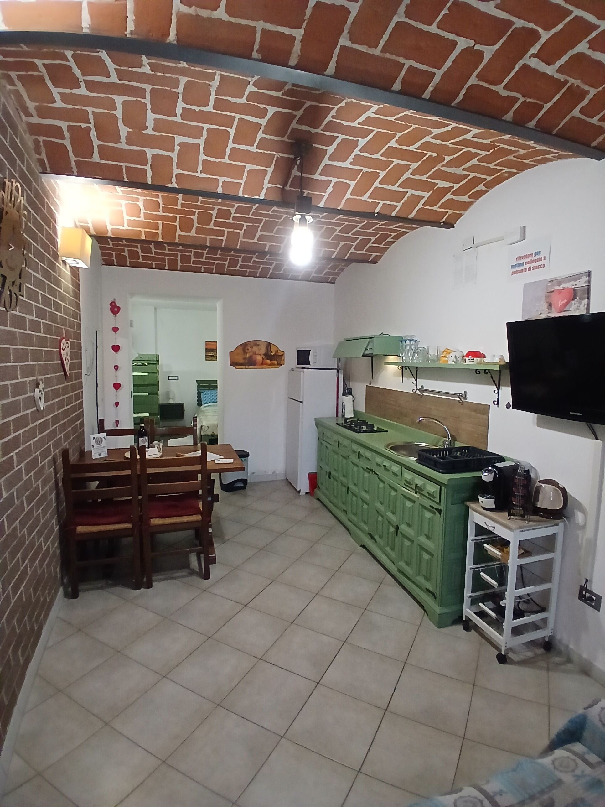 A kitchen with a brick ceiling, green cabinets, a dining table with chairs, and a television mounted on the wall.
