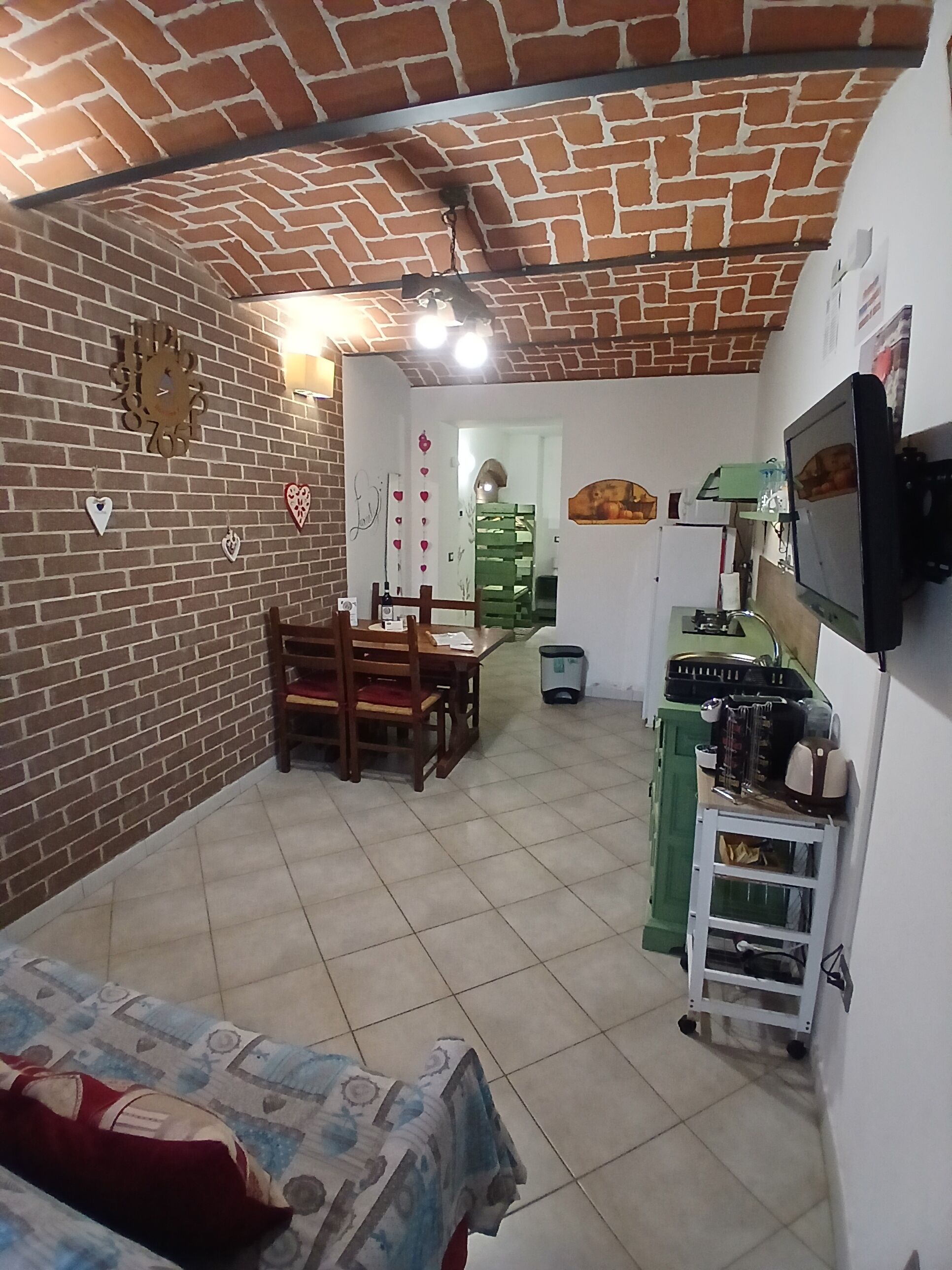 A cozy room with a brick wall, a wooden dining table, a TV mounted on the wall, and a green cart with various items.
