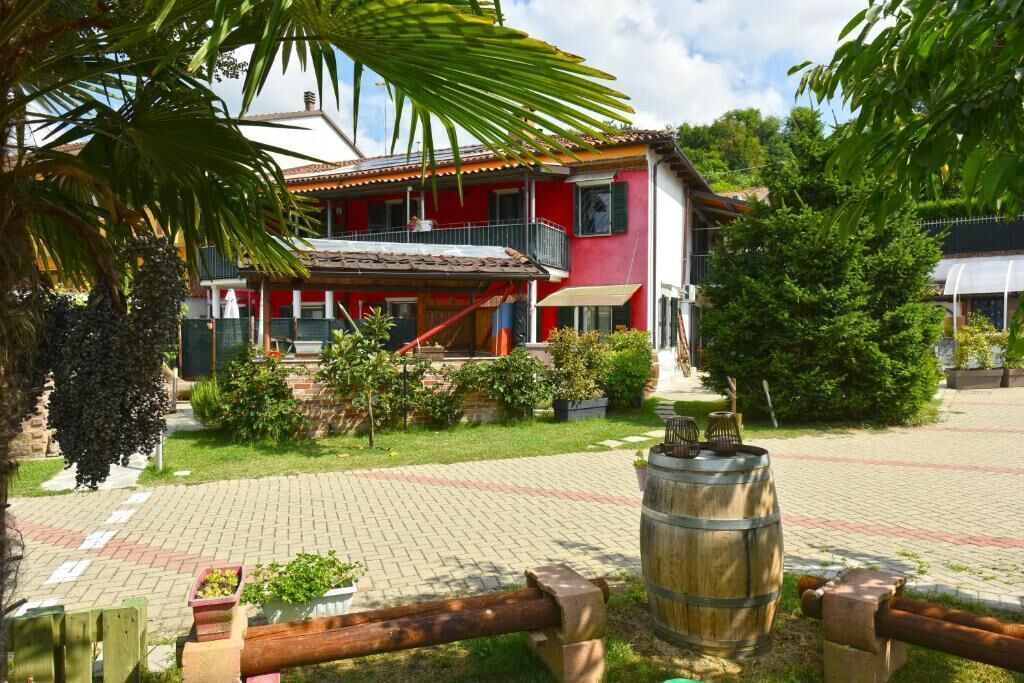 A red building with a balcony, surrounded by greenery and a paved area.