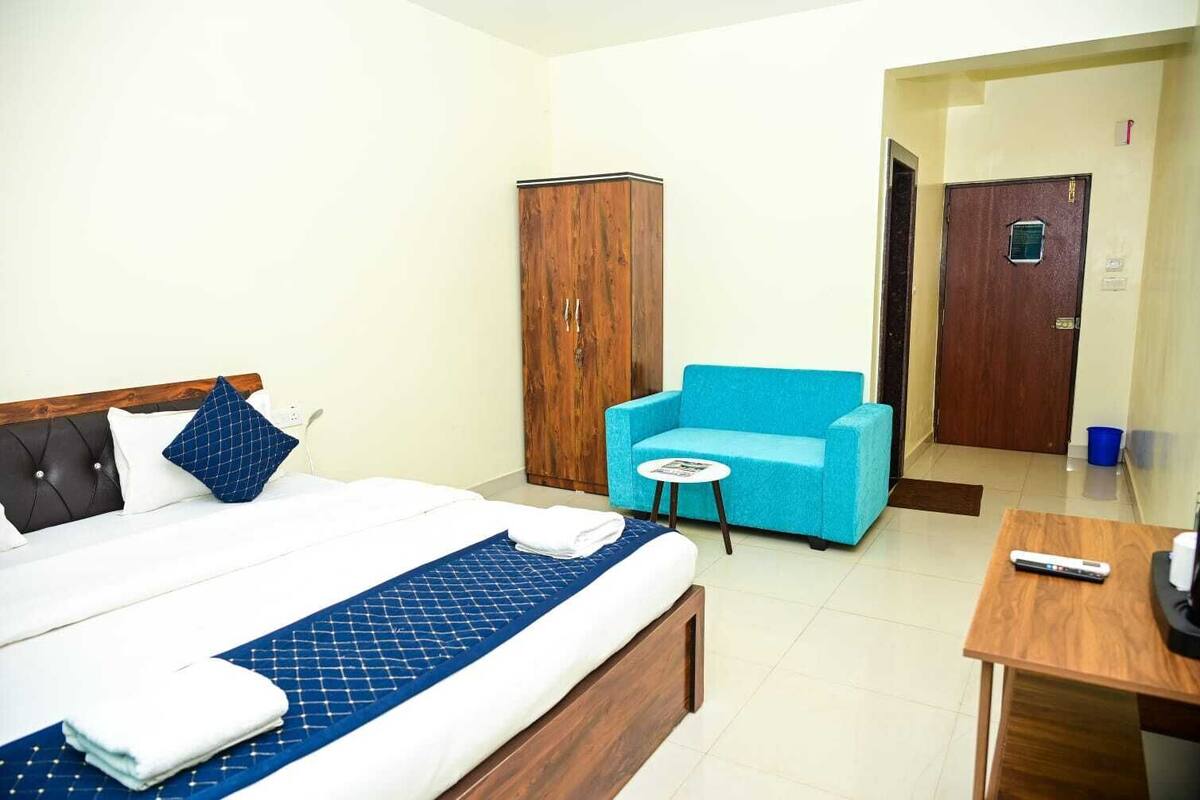 Habitación de hotel con cama, un sillón azul, un armario de madera, una mesita pequeña y un escritorio con control remoto.