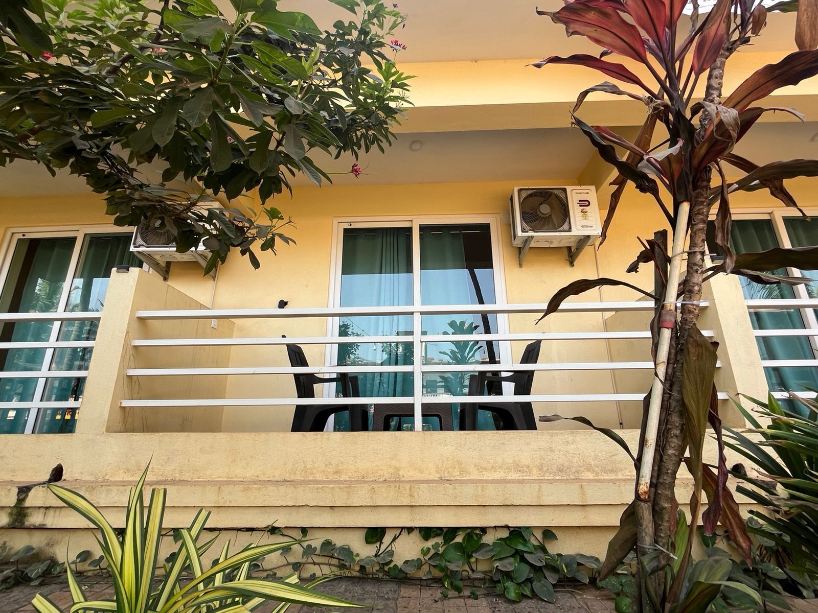 Un bâtiment à la façade jaune, doté de climatiseurs, d’un balcon avec une balustrade blanche et agrémenté de diverses plantes.