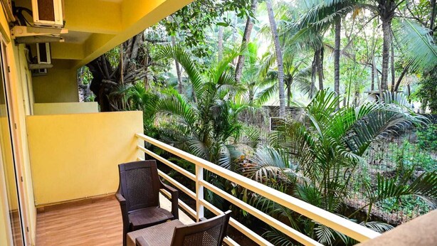 Un balcón con suelo de madera, dos sillas de mimbre y barandilla amarilla, con vistas a un jardín exuberante y plantas tropicales diversas.