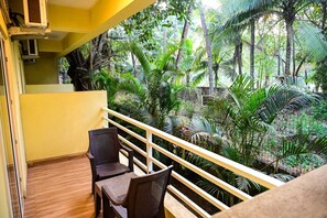 A balcony with a wooden floor, two wicker chairs, and a yellow railing overlooking a lush garden with various tropical plants. - Navratna Guest House (Morjim)