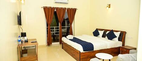 A hotel room with a bed, a desk, a chair, a TV, and an air conditioning unit.