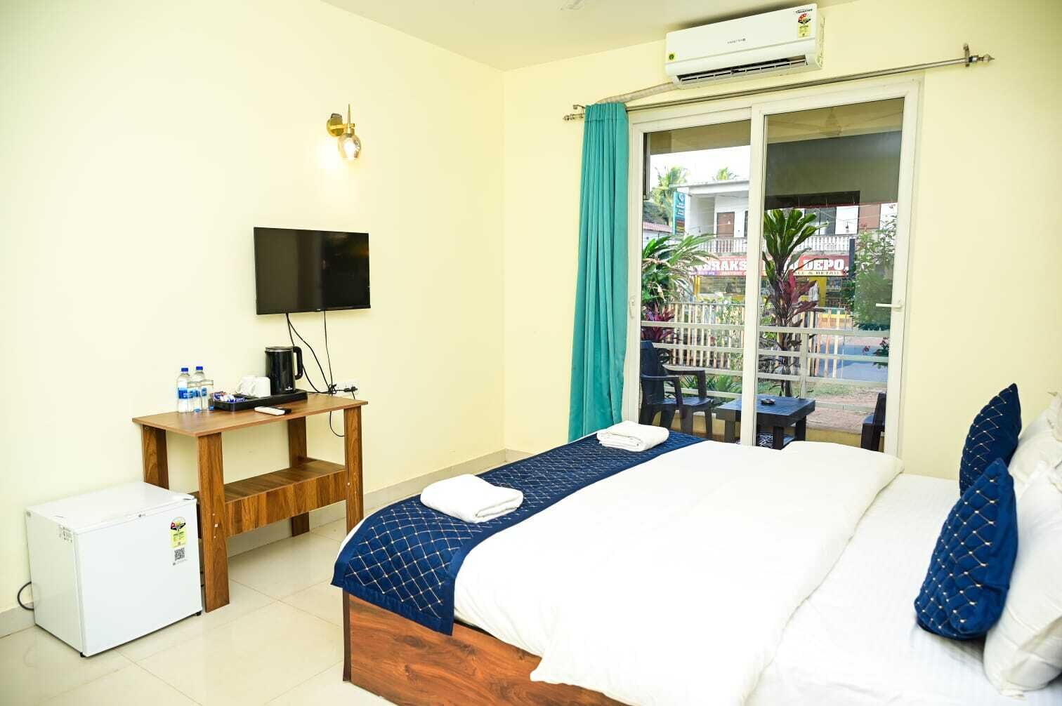 A hotel room with a bed, a desk with a TV, a mini-fridge, and a balcony with a view of the outside.