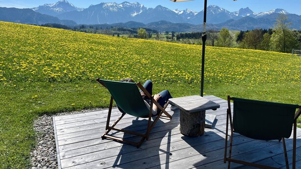 A deck with two chairs and a table, an umbrella, and a field of yellow flowers with mountains in the background. - Vacation home Benedikt Füssen, Eschach FW2 (Füssen)