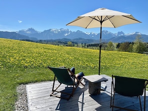Une terrasse avec deux chaises et une table, un parasol, et un champ de fleurs jaunes avec des montagnes en arrière-plan.