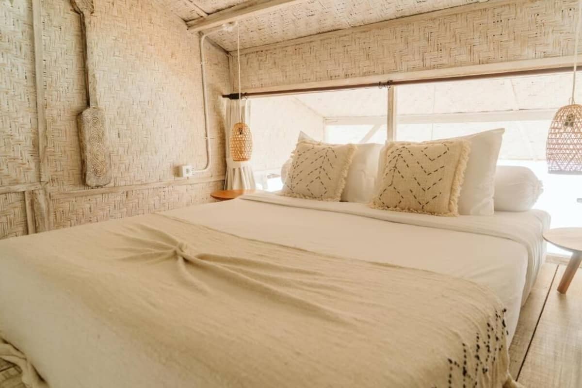 A bed with a white bedspread and pillows, a woven wall, and a hanging light fixture.