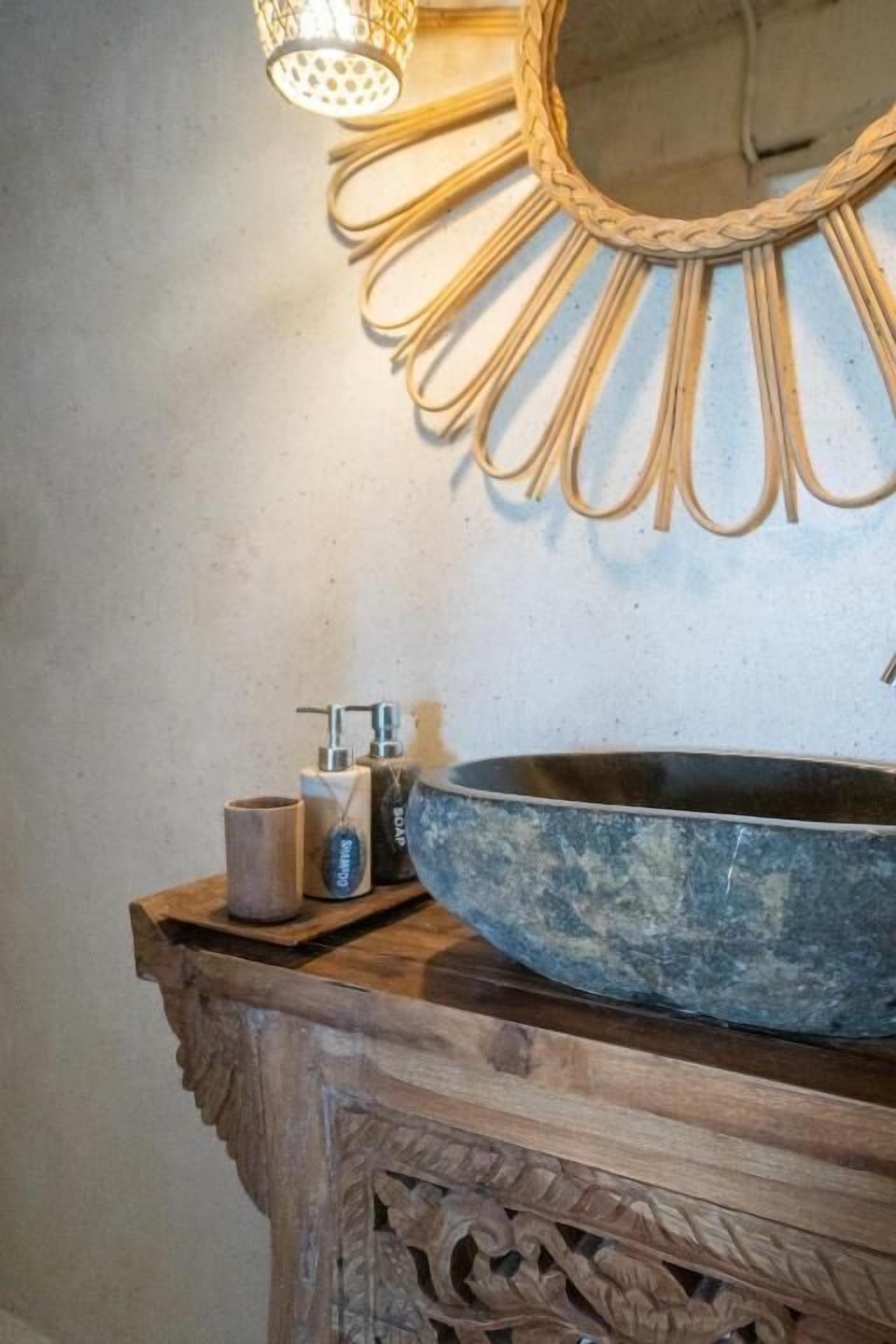 A bathroom with a round, dark-colored basin, a wooden vanity with intricate carvings, a wall-mounted mirror with a sunburst design, and a small, textured container on the vanity.