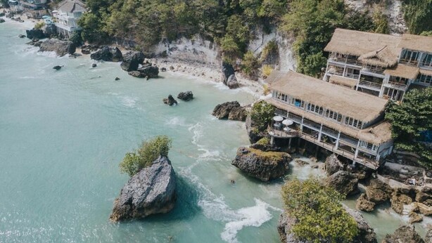 A coastal hotel with multiple buildings, including a thatched-roof structure with outdoor seating, situated on rocky terrain with clear turquoise waters. - Dreamsea Uluwatu (Pecatu)