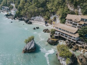 Um hotel costeiro com vários edifícios, incluindo uma construção com telhado de palha e área de estar ao ar livre, localizado em terreno rochoso com águas turquesa cristalinas.