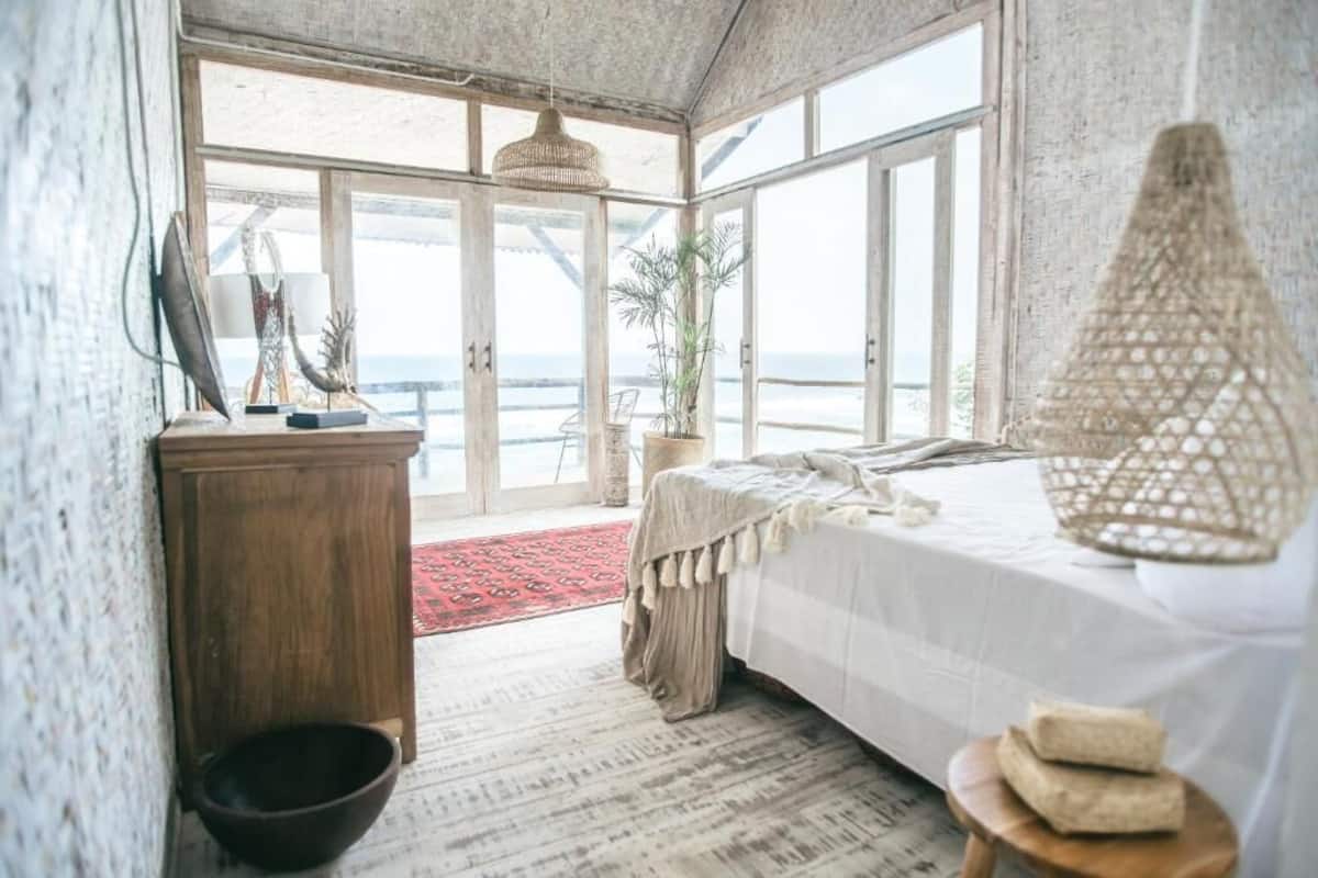 A bedroom with a large window, a bed, a wooden dresser, a hanging light fixture, and a red rug.