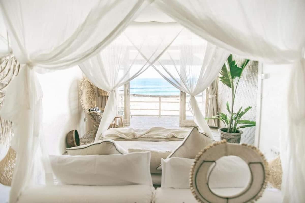 A room with a bed, white curtains, a view of the beach, and a potted plant.