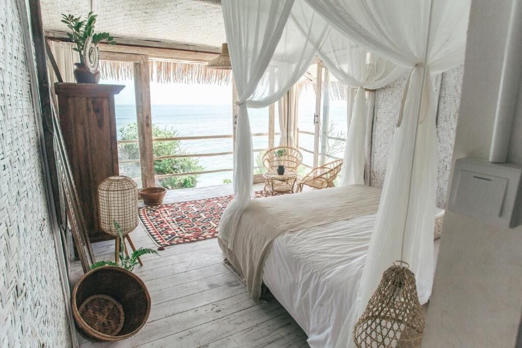 A bedroom with a bed, a wicker chair, a small table, and a view of the ocean.