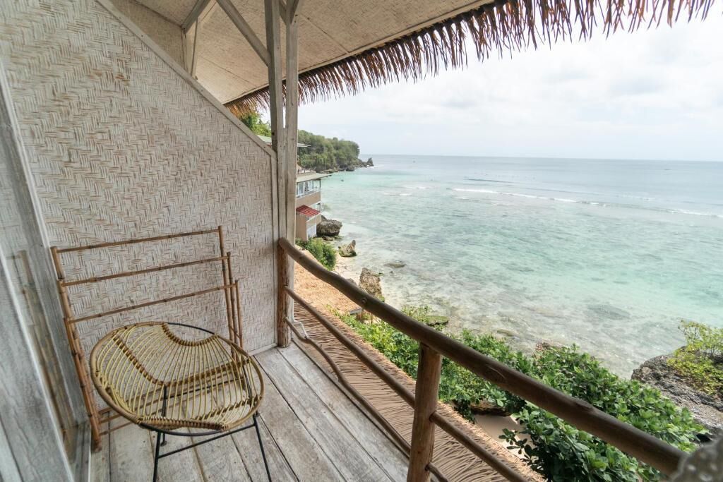 A wooden deck with a wicker chair overlooking a beach with clear turquoise water and rocky shoreline.