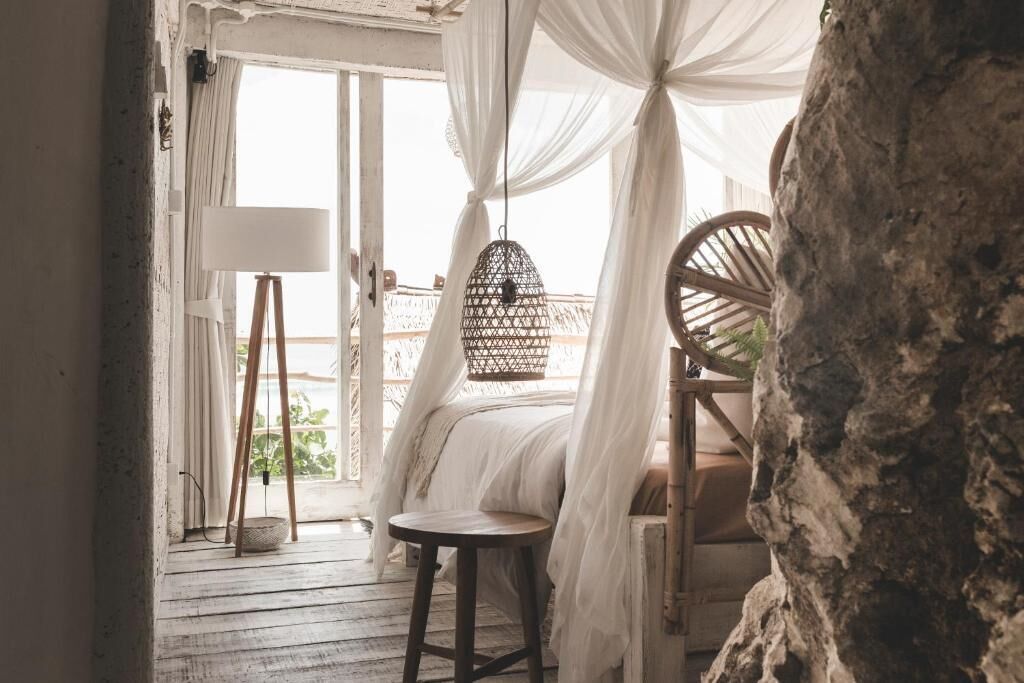 Una habitación con una cama bajo un dosel, una silla de mimbre, una mesita redonda y una gran roca.