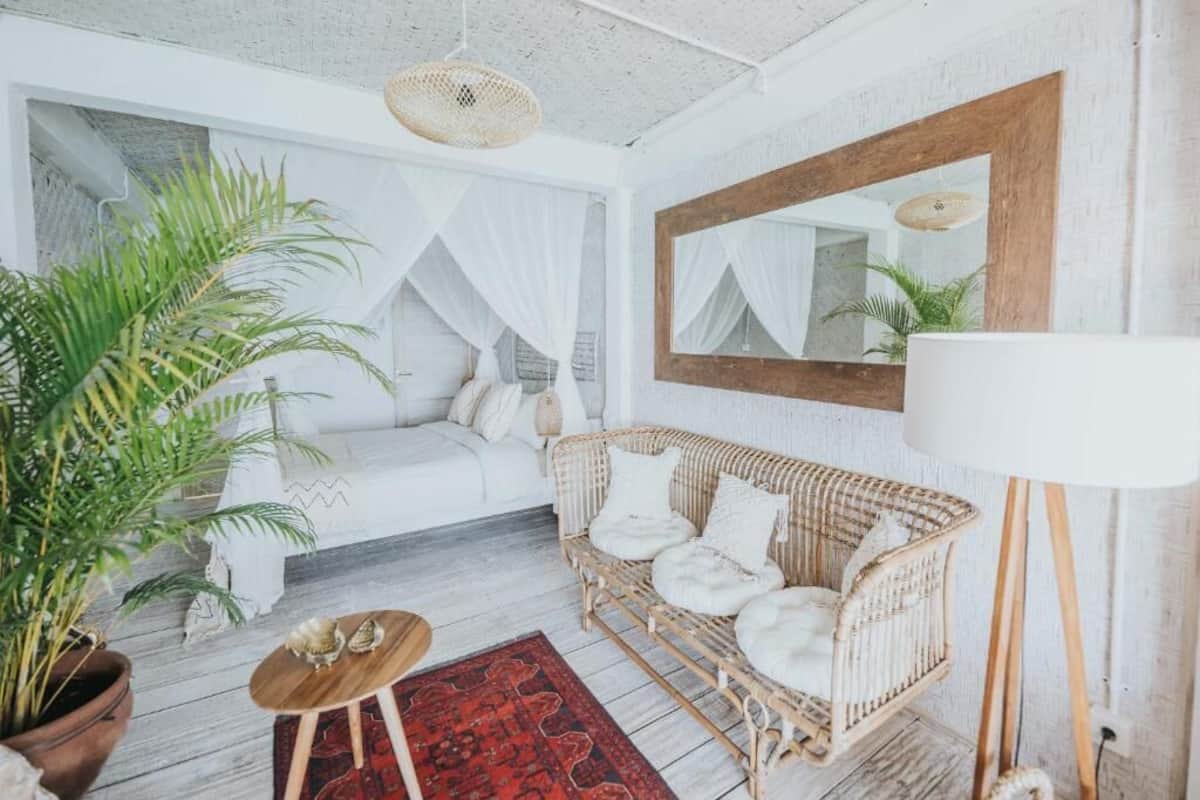 A cozy room with a white bed, a wicker sofa, a large mirror, and a potted plant.