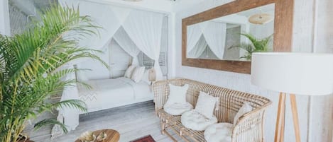 A cozy room with a white bed, a wicker sofa, a large mirror, and a potted plant.