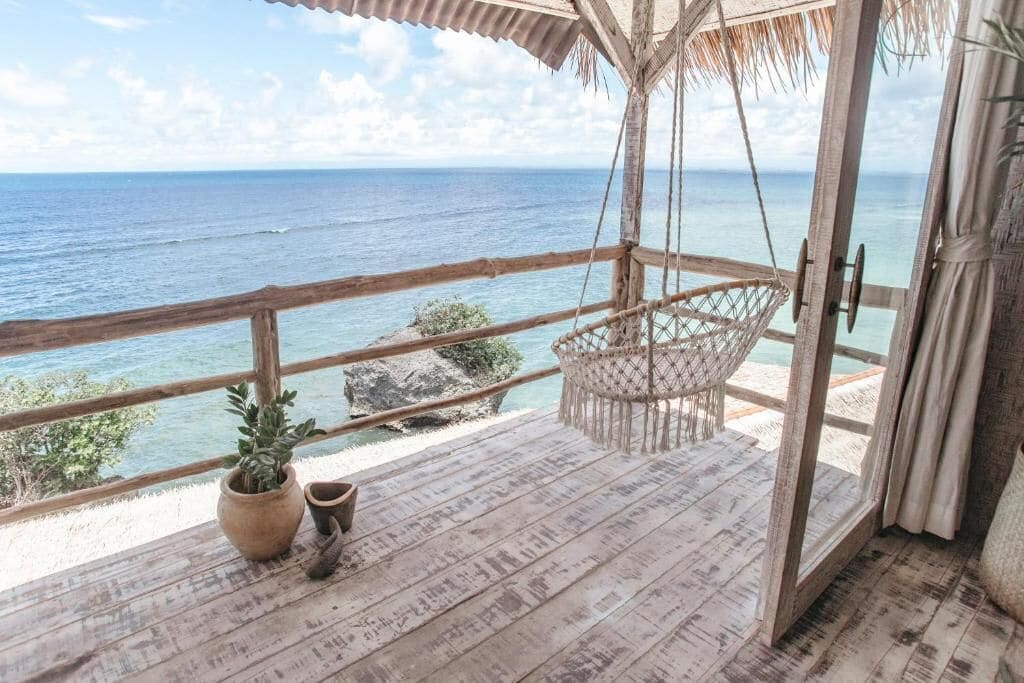 Una terraza de madera con una silla colgante, una planta en maceta y vistas al océano.