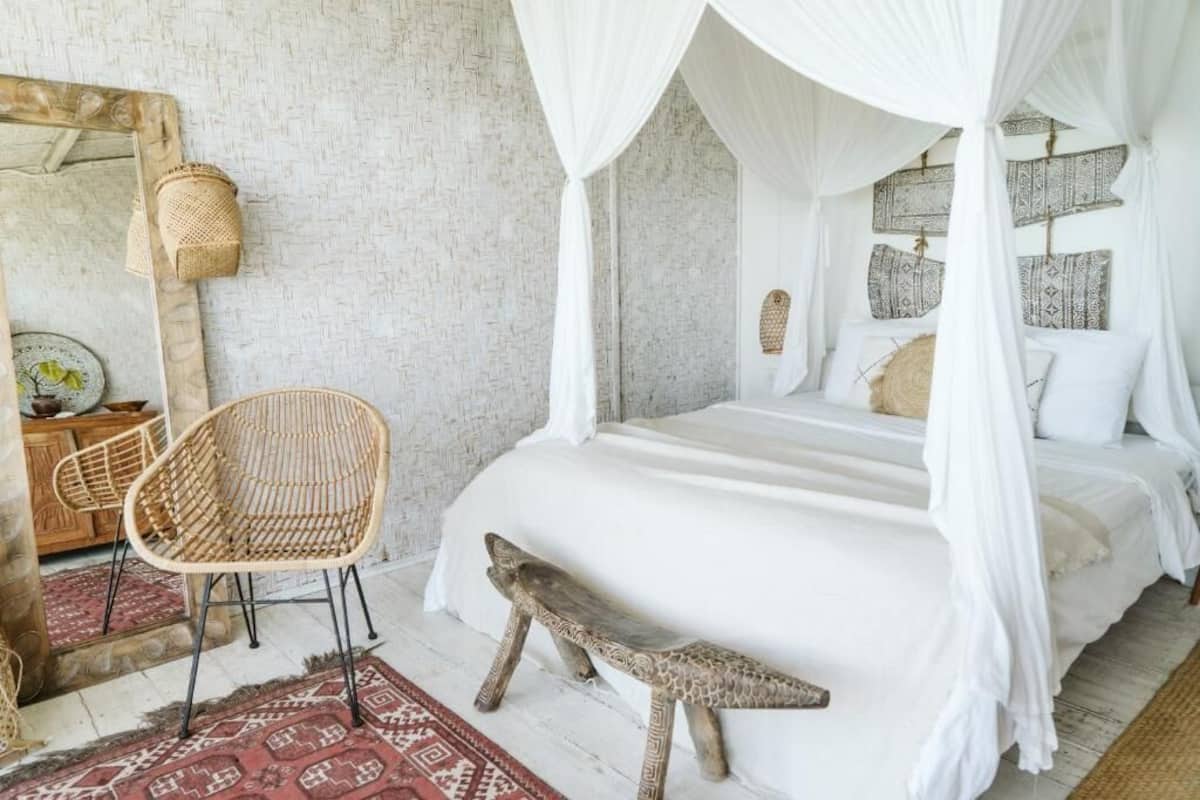 A bed with a canopy, a wicker chair, a bench, a mirror, and patterned cushions.