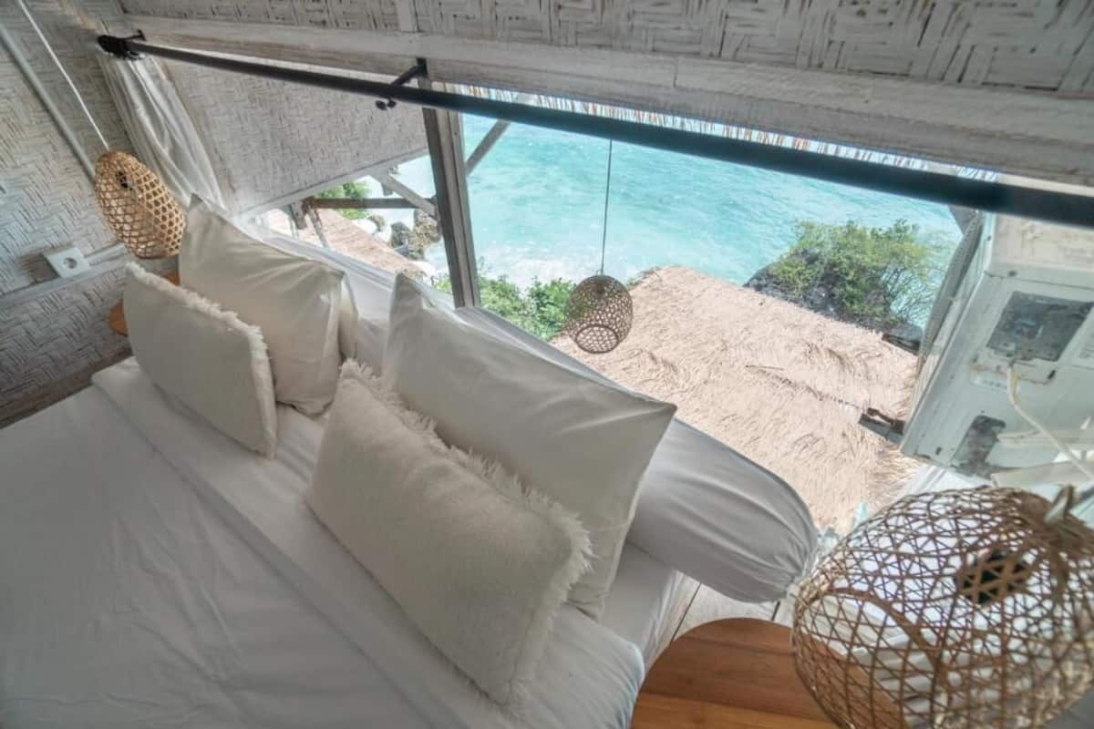 A bed with white linens and pillows, a view of the ocean through a window, and a woven light fixture.