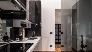 A modern kitchen with a black stove, white countertops, and a wooden floor. There is a black bookshelf in the background. - Giotto (Matraia)
