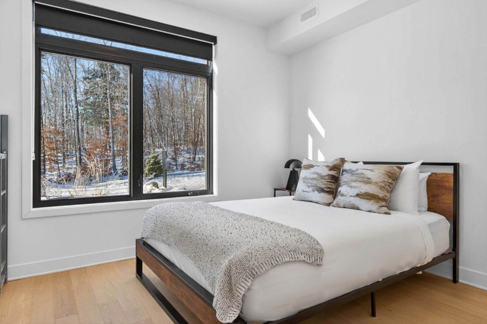 A bedroom with a large window, a bed with a white bedspread and two pillows, a wooden headboard, and a floor lamp.