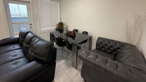 A modern living room with a black leather sofa, a dining table with chairs, and a large window with blinds. - A home away from home. YEG retreat (Edmonton)