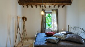 Ein Schlafzimmer mit Bett, einer Hängelampe, einem Fenster mit Vorhängen und einer Holzbalkendecke.