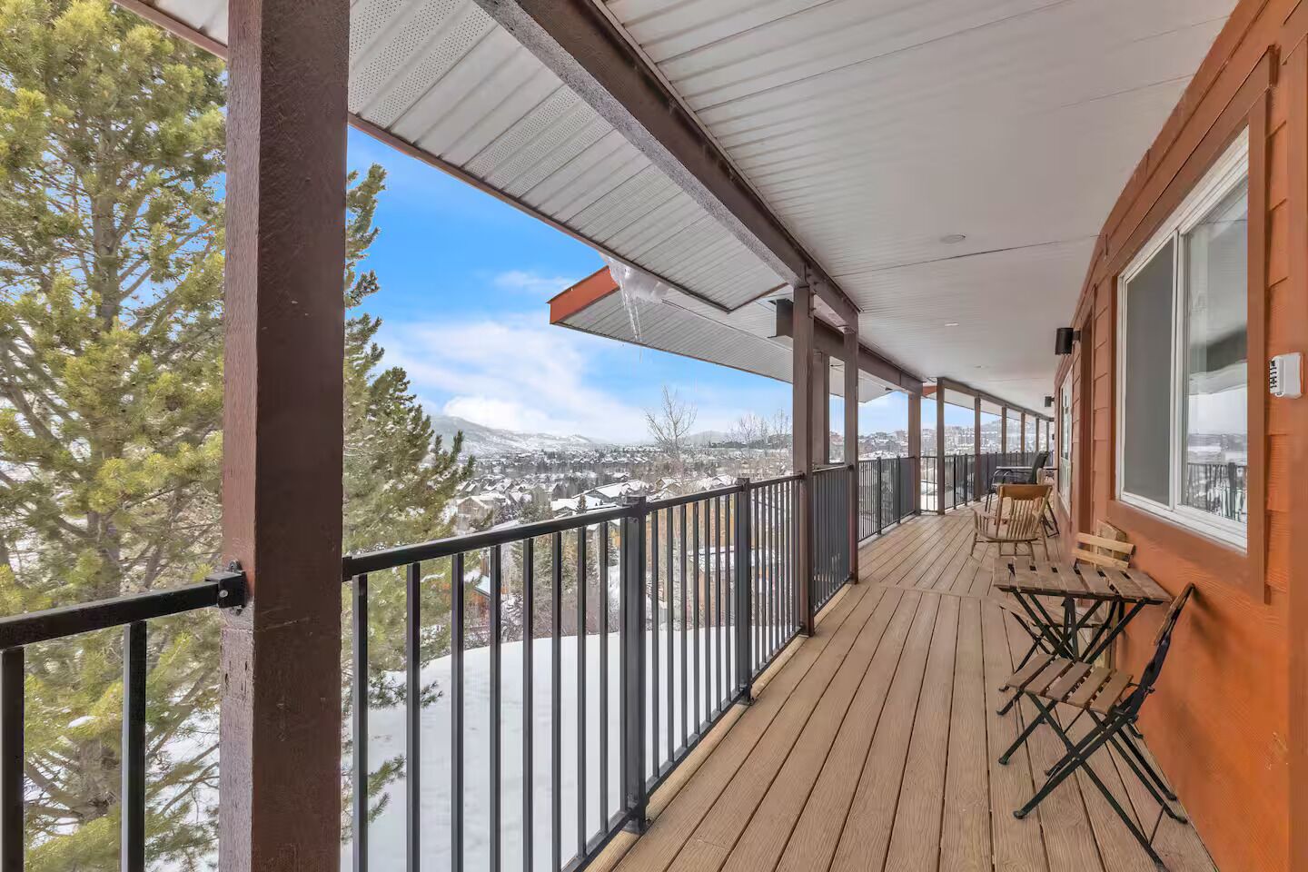 A wooden deck with a metal railing, outdoor seating, and a view of a snowy landscape.