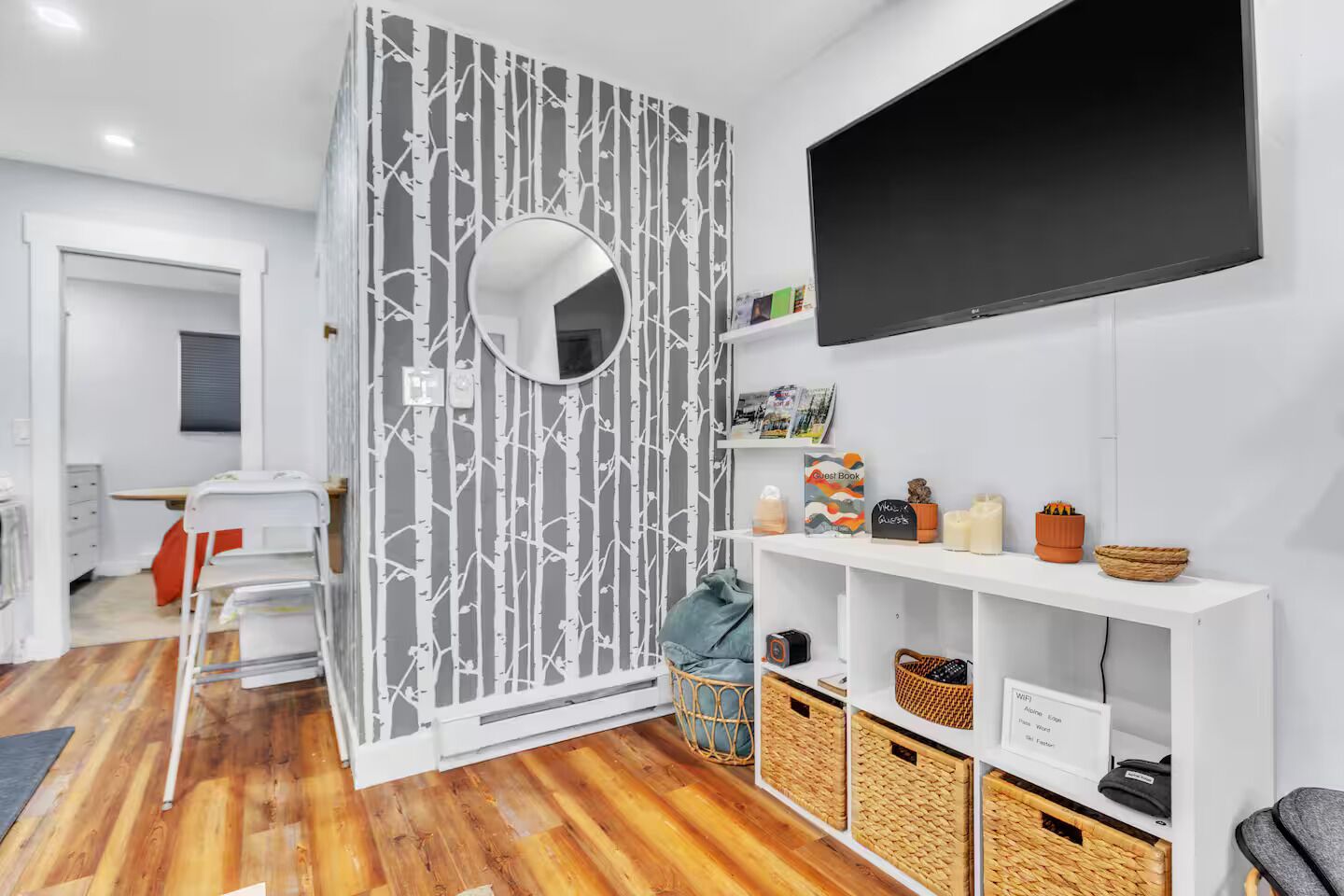 Una camera d'albergo moderna con pavimento in legno, un armadio bianco, una TV a parete e una piccola area salotto con una sedia e un tavolo.