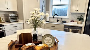 A kitchen with white cabinets, a stove, a sink, and a window with a view. There is a wooden tray with two loaves of bread and a vase with white flowers. - Scenic Sky Retreat | EV Charger & Skyline Views (Chattanooga)