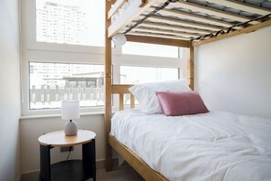 Een stapelbed met wit beddengoed en een roze kussen, een nachtkastje met een lamp, en een raam met uitzicht over de stad.