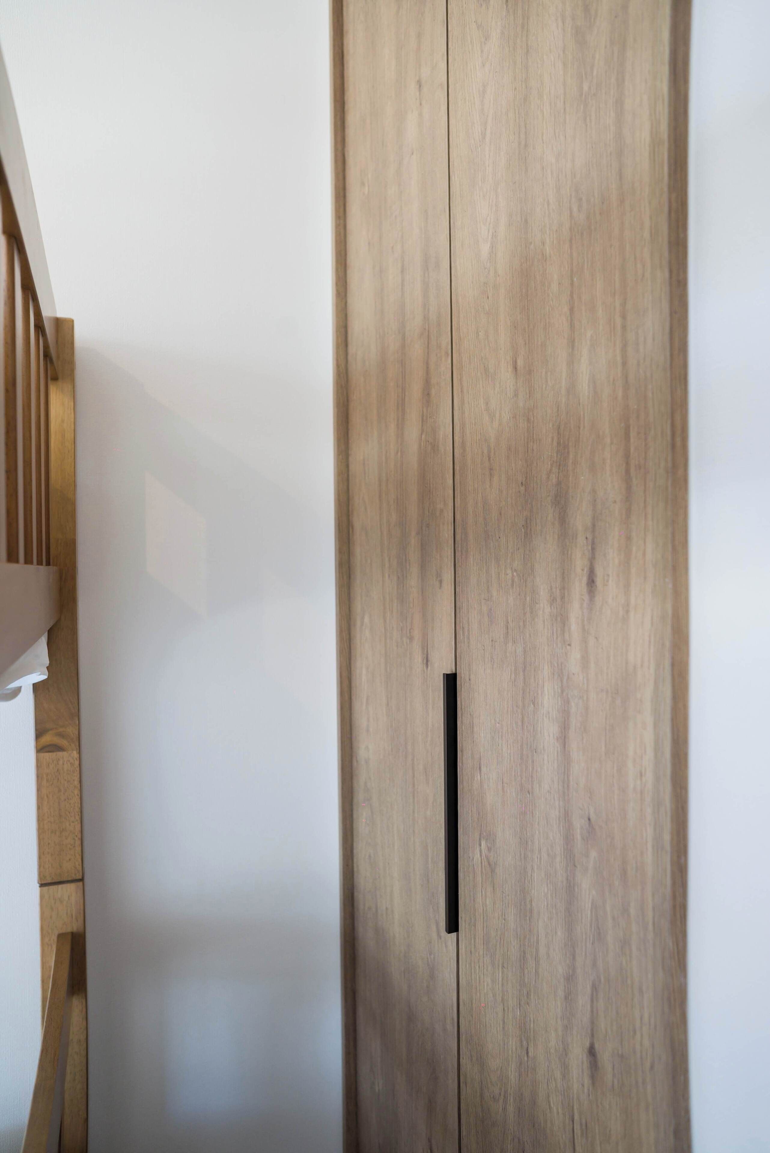 A wooden wardrobe with a black handle against a plain wall.